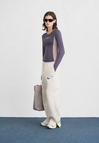 Μπλούζα με μακριά μανίκια σε μωβ χρώμα με ανοιχτό γκρι πλευρικό πάνελ, λευκό παντελόνι με κορδόνι και μπλε λογότυπο, και γκρι τσάντα tote. Συνοδεύεται από γυαλιά ηλίου.