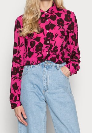Frau mit langen blonden Haaren trägt eine leuchtend pinke Bluse mit schwarzem Blumenmuster und hellblaue High-Waist-Jeans.