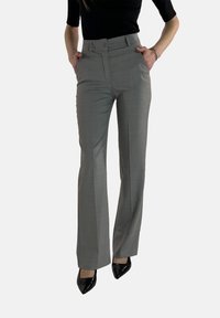 Pantalon tailleur gris avec une taille haute, un devant plat et des poches latérales. Le tissu a une texture subtile, associé à des talons noirs brillants.
