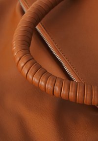 Poignée de sac en cuir marron avec texture striée. Piste de fermeture éclair en métal visible sur le corps en cuir marron doux, soulignant un design harmonieux.