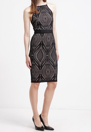 Mujer con vestido negro sin mangas hasta la rodilla con patrón geométrico de encaje y zapatos negros de tacón alto, posando contra un fondo blanco.