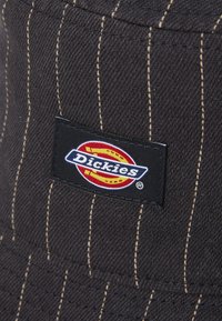 Schwarzer Bucket Hat mit vertikalen Nadelstreifen, ausgestattet mit einem auffälligen weißen und roten "Dickies"-Logo-Patch. Strukturierter Stoff.