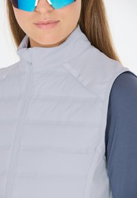 Ljust blå ärmlös väst med hög krage, som har quiltade paneler och en mjuk textur, bärs över en mörkblå långärmad skjorta.