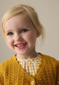 Keltainen neulottu villatakki, jossa on aaltoilevat reunat, teksturoitu kuvio ja puinen nappi, päällä valko-keltainen ruutupaitakuvio.