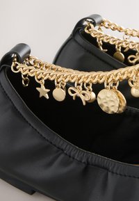 Chaussure plate en cuir noir avec bride chaîne dorée ornée d'étoile, cercle, nœud et breloques rondes texturées.