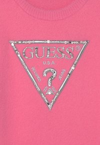 Maglione in pile rosa con un logo triangolare in argento metallizzato con la scritta "GUESS U.S.A." e decorazioni lungo i bordi.