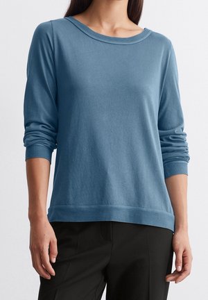Femme portant un pull bleu à manches longues avec poignets et ourlet côtelés, associé à un pantalon noir, visible du cou aux hanches.