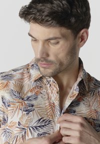 Joven con cabello corto y oscuro, vistiendo una camisa beige con patrones de hojas en naranja, marrón y negro, abotonándose la camisa.