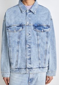 Giacca di denim azzurro chiaro con due tasche sul petto, bottoni argentati e un'etichetta con logo. Design classico con una vestibilità rilassata e cuciture visibili.
