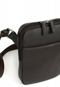 Borsa a tracolla in pelle marrone scuro con una superficie liscia, chiusura a zip e tasca frontale. Caratterizzata da una tracolla regolabile e hardware minimal.