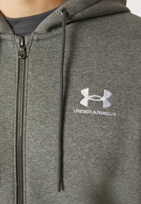 Felpa con zip grigia realizzata in tessuto morbido, dotata di due lacci, con il logo Under Armour ricamato in bianco sul lato sinistro del petto.