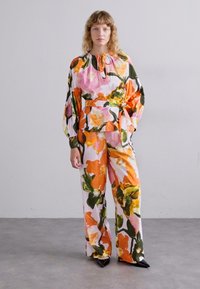 Top con collo alto a fantasia floreale con maniche arricciate abbinato a pantaloni ampi. I colori includono rosa, arancione e verde su uno sfondo bianco.