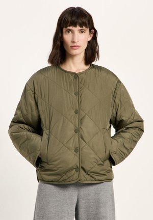 Frau trägt eine olivgrüne gesteppte Jacke mit Druckknöpfen und den Händen in den Taschen, steht vor einem schlichten hellen Hintergrund.