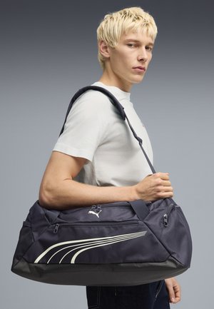 Giovane uomo con una t-shirt bianca che porta una borsa da palestra Puma blu navy a tracolla su uno sfondo grigio semplice.