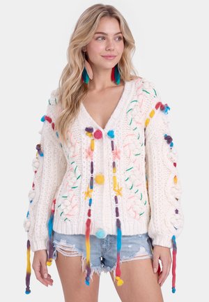 Ung kvinde med langt blondt hår bærer en hvid strikket sweater dekoreret med farverige garnudsmykninger og slidte denimshorts.