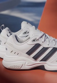Paire de baskets blanches Adidas avec des bandes noires reposant sur une surface orange, montrant les détails latéraux et supérieurs.