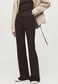 Personne portant un pantalon large marron foncé, un haut côtelé marron foncé, une veste beige et des chaussures blanches pointues, se tenant avec une main dans la poche de la veste.