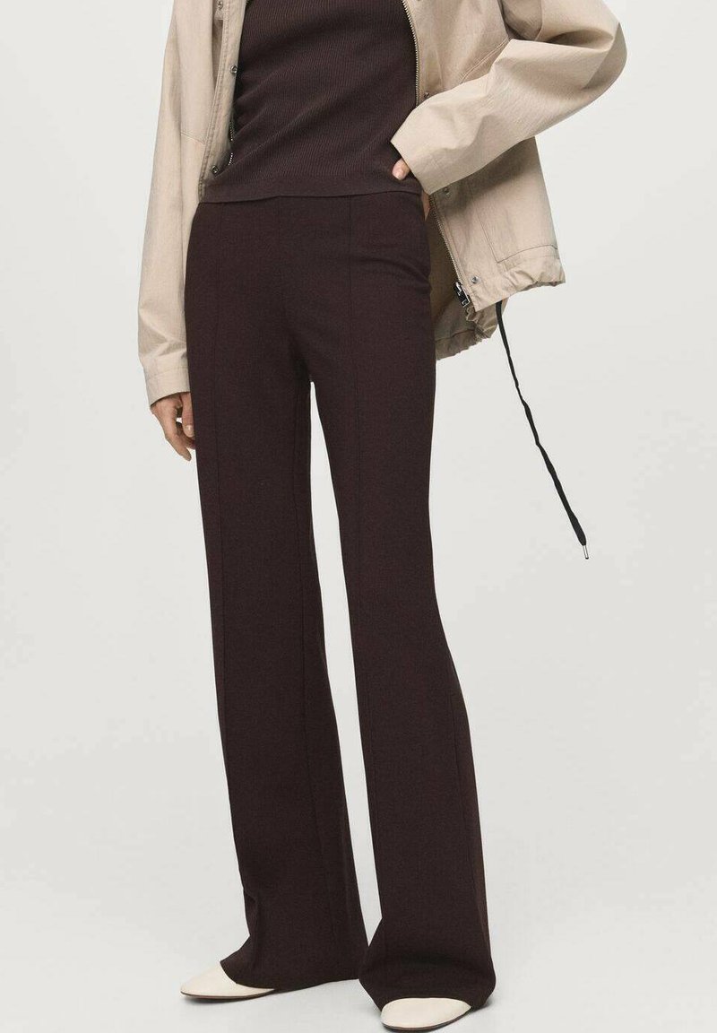 Personne portant un pantalon large marron foncé, un haut côtelé marron foncé, une veste beige et des chaussures blanches pointues, se tenant avec une main dans la poche de la veste.