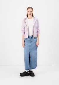 Giacca leggera zip-up lavanda, t-shirt bianca, gonna maxi in denim blu con spacco anteriore e scarpe platform nere. Outfit casual.