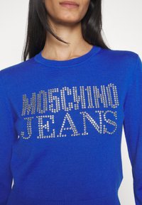 Femme portant un pull bleu vif avec "MOSCHINO JEANS" écrit en clous argentés sur le devant.