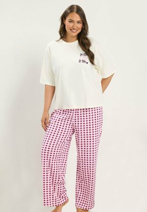Mujer sonriente con el cabello largo y oscuro, que lleva una camiseta blanca holgada y pantalones sueltos a cuadros rosas y morados, de pie frente a un fondo liso.