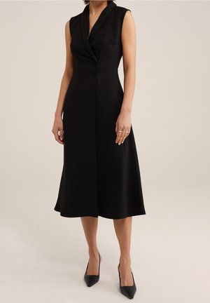 Ärmelloses schwarzes Midi-Kleid mit Reverskragen und seitlichen Knöpfen, kombiniert mit schwarzen spitzen Pumps vor neutralem Hintergrund.