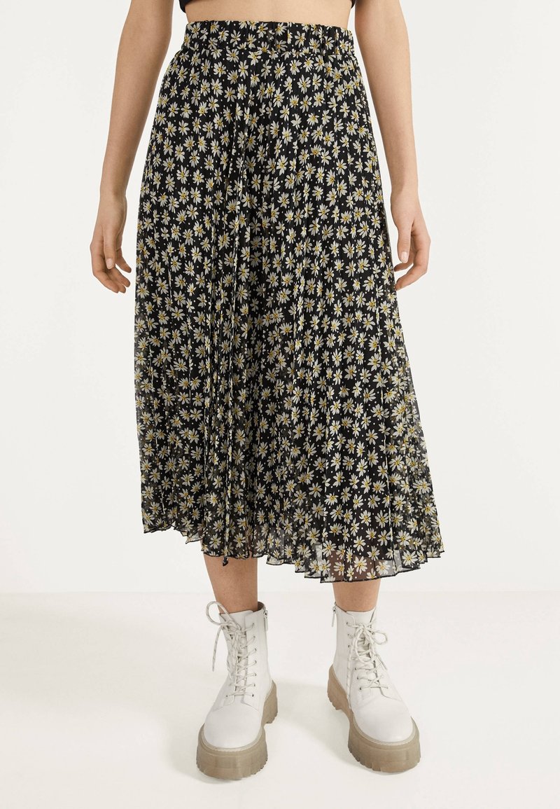 Falda midi plizada negra con un patrón de margaritas en blanco y amarillo, con una cinturilla elástica y una textura fluida, acompañada de botas blancas.