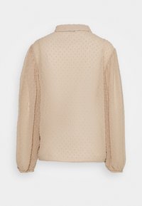 Blouse beige avec des pois texturés, manches longues ballons avec poignets froncés et un col classique. La vue de dos montre une finition lisse.