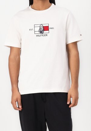 Osoba ubrana w białą koszulkę z krótkim rękawem z nadrukiem łodzi i napisem „EST. 1985 HILFIGER” na klatce piersiowej, w połączeniu z czarnymi spodniami.