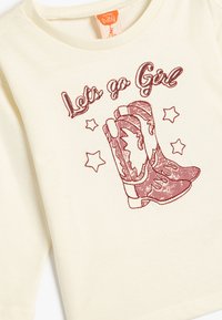 Cremefarbene Baumwoll-T-Shirt mit einem Grafikmotiv von Cowboy-Stiefeln in Bordeauxrot, umgeben von Sternen. Der Text lautet: "Lass uns gehen, Mädchen." Rundhals und lange Ärmel.