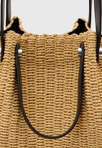 Borsa a tracolla in paglia intrecciata con manici in pelle nera e una barra metallica infilata nei passanti dei manici vicino al bordo superiore della borsa.