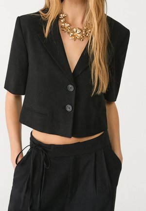 Vrouw met een cropped zwarte blazer met twee knopen, een zwarte broek met hoge taille en trekkoord, en een gouden bloemige ketting.