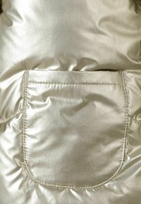 Section de blouson rembourré en or métallisé présentant un tissu brillant, une texture matelassée et une poche cousue. Finition lisse avec des coutures visibles.