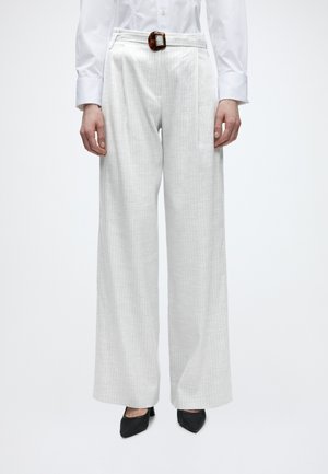 Person iført hvide vidde bukser med smalle striber og brunt bælte, hvid langærmet skjorte og sorte spidse hæle, stående oprejst.