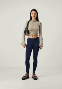 Moteris stovi vilkėdama trumpą ilgomis rankovėmis smėlio spalvos marškinėlį, tamsiai mėlynas aptemptas džinsus, juodas šlepetes ir juodą rankinę ant peties.