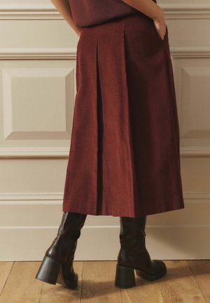 Femeie purtând o fustă midi plisată de culoare ruginie și cizme negre din piele cu toc, stând pe podea din lemn, cu un perete cu panouri pe fundal.