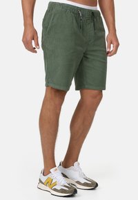 Grüne Cordshorts mit elastischem Bund und Kordelzug, ausgestattet mit zwei Seitentaschen und einem lässigen Schnitt, getragen mit weißen und grauen Sneakers.