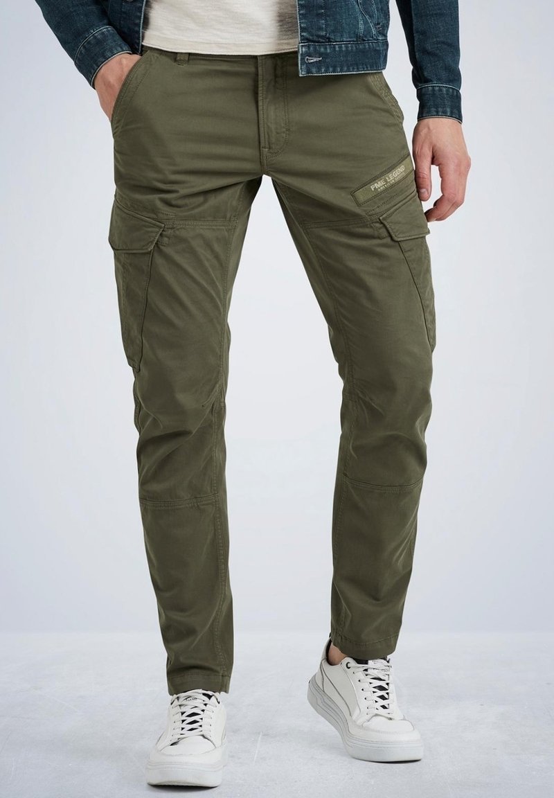 Homme portant un pantalon cargo vert olive avec des poches latérales, des baskets blanches, une chemise blanche et une veste en jean foncée.