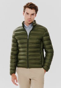 Oliwkowa kurtka puchowa z pikowanym wzorem i kołnierzem stojącym. Posiada zamek z przodu oraz kieszenie po bokach, noszona na wzorzystej koszuli.