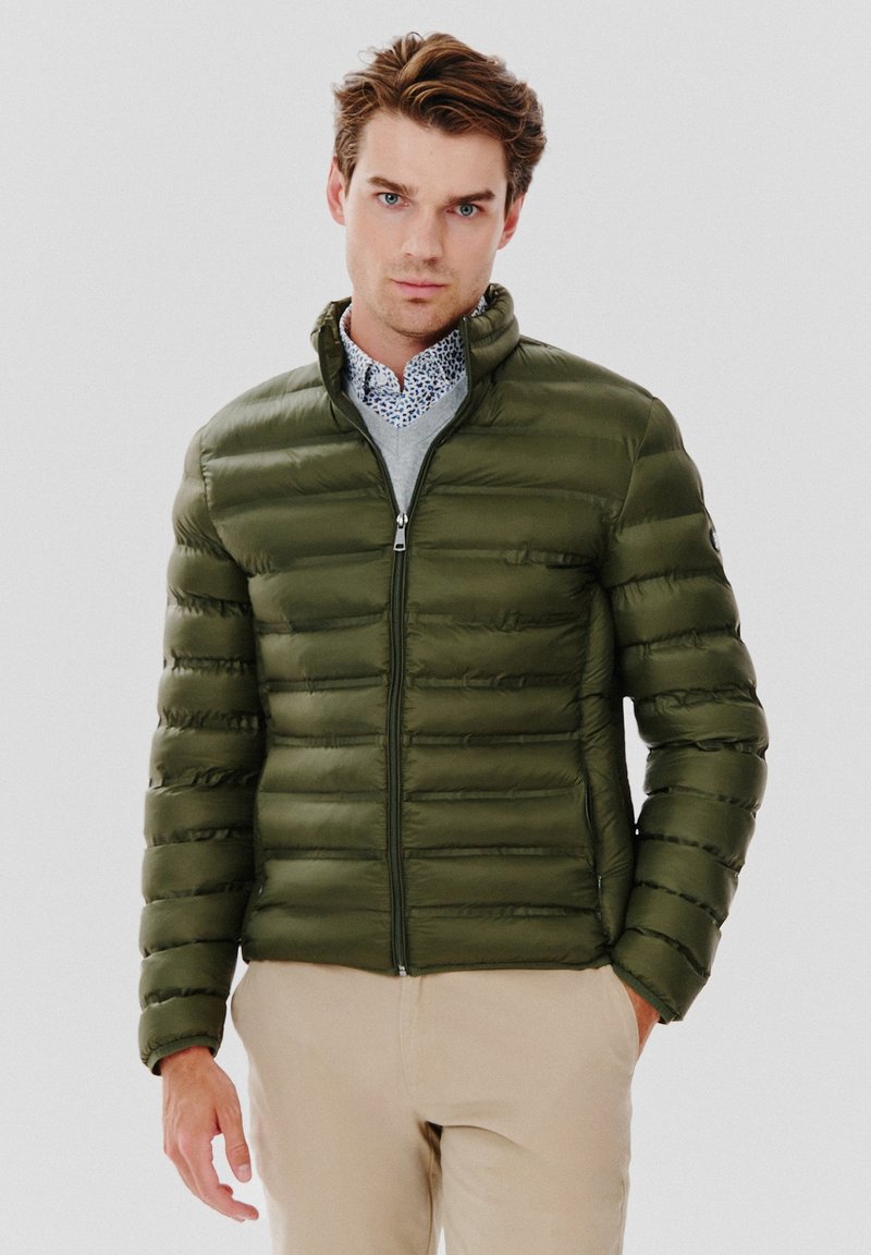 Oliwkowa kurtka puchowa z pikowanym wzorem i kołnierzem stojącym. Posiada zamek z przodu oraz kieszenie po bokach, noszona na wzorzystej koszuli.