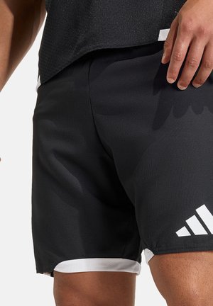 Sportowiec ubrany w czarne i białe spodenki sportowe Adidas oraz czarną teksturowaną sportową koszulkę, stojący z ręką swobodnie opuszczoną wzdłuż ciała.