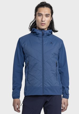 Mann mit langen Haaren, der eine blaue, zip-up Kapuzenjacke und dunkle Hosen trägt, vor einem schlichten hellen Hintergrund stehend.