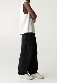 Persona de piel oscura con una camiseta sin mangas blanca, pantalones negros sueltos y zapatillas blancas, de pie con los brazos cruzados frente a un fondo liso.