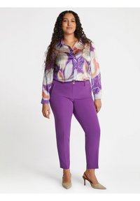 Blusa a maniche lunghe bianca con un motivo floreale astratto in tonalità di viola e pesca, abbinata a pantaloni viola a gamba stretta e tacchi beige.