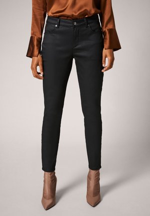 Femme portant un pantalon noir ajusté, un chemisier en satin marron avec des manches évasées et des bottines marron à bout pointu, debout devant un fond uni.