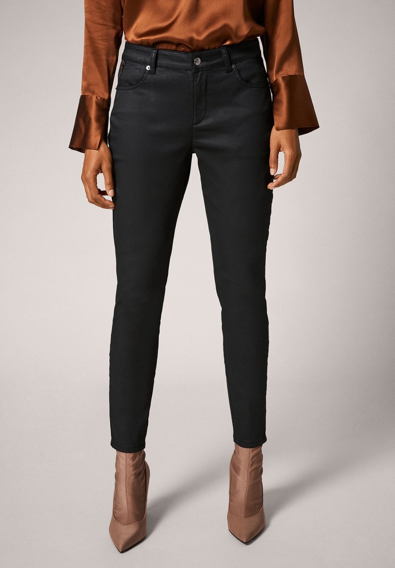 Femme portant un pantalon noir ajusté, un chemisier en satin marron avec des manches évasées et des bottines marron à bout pointu, debout devant un fond uni.
