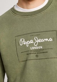 Acercamiento a una sudadera verde con el logo y texto en blanco y negro de "Pepe Jeans Londres" en el pecho.
