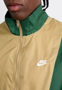 Nike jakna z visokim ovratnikom, z bež telesom in zelenimi detajli. Opozorljiv bel logo na prsih in zadrgo spredaj. Gladka tekstura.