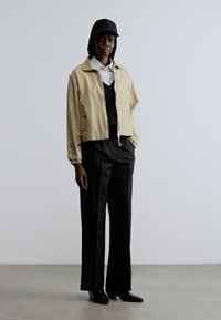 Lichtbeige bomberjack over een zwarte top, gecombineerd met zwarte wijde broekspijpen. Zwarte pet en glanzende zwarte schoenen maken de outfit compleet.