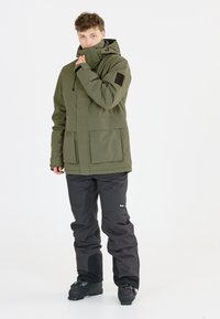 Olivgrüne isolierte Jacke mit hohem Kragen, großen Fronttaschen und verstellbarem Kapuzen, kombiniert mit schwarzen wasserdichten Hosen und Stiefeln.
